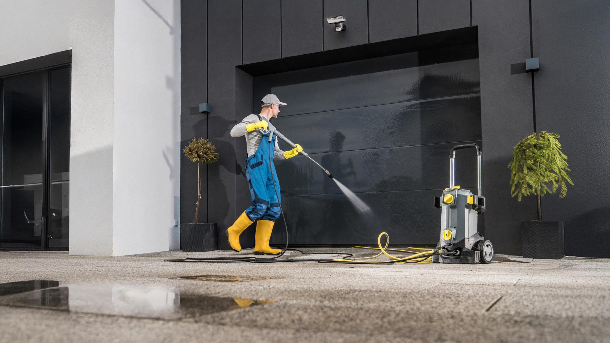 A person in blue overalls and yellow boots uses a pressure washer to clean a garage door, with a water hose connected to the machine. The setting features a modern building, and there are potted plants nearby.